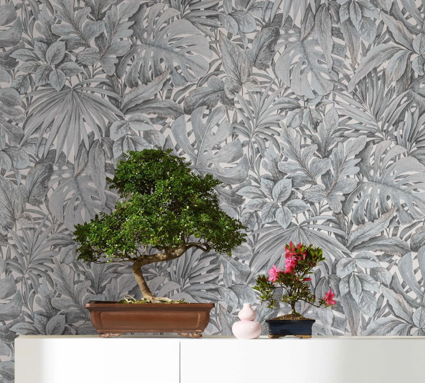 Marburg Botanica 33306 Gray tropical leaf wallpaper serves as the backdrop for two bonsai plants on a white cabinet, with a small pink vase beside them.