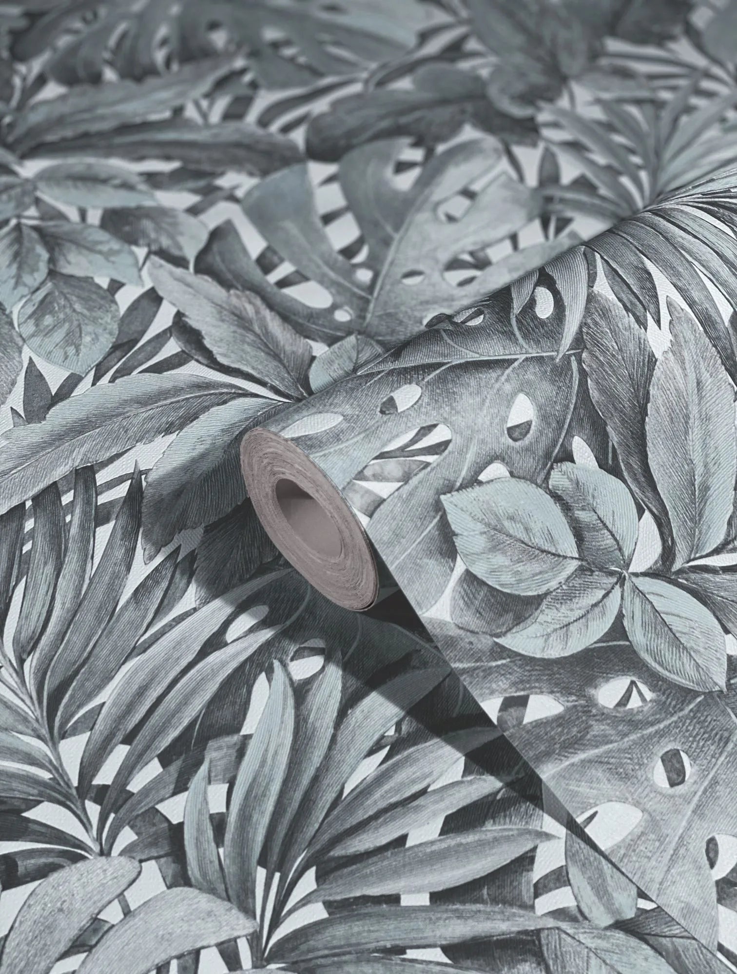 Marburg Botanica 33306 Close-up of a wallpaper roll resting on matching paper with a dense tropical leaf pattern, showing layered palm and split-leaf foliage in muted green tones on a light background.