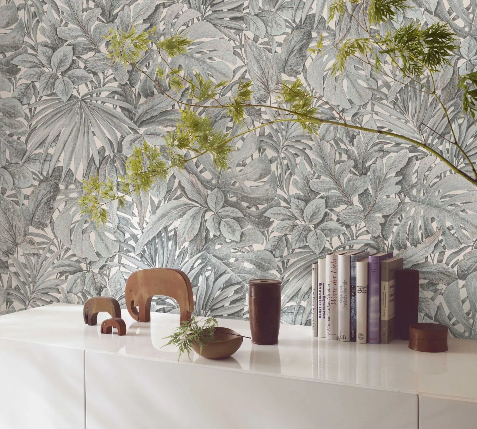 Marburg Botanica 33306 Botanical wallpaper with dense overlapping tropical leaves in muted green and white behind a white sideboard styled with books, small elephant figurines, a shallow bowl, a dark cylindrical cup, a small lidded container, and a leafy branch extending across the.