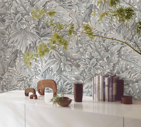 Marburg Botanica 33306 Botanical wallpaper with dense overlapping tropical leaves in muted green and white behind a white sideboard styled with books, small elephant figurines, a shallow bowl, a dark cylindrical cup, a small lidded container, and a leafy branch extending across the.