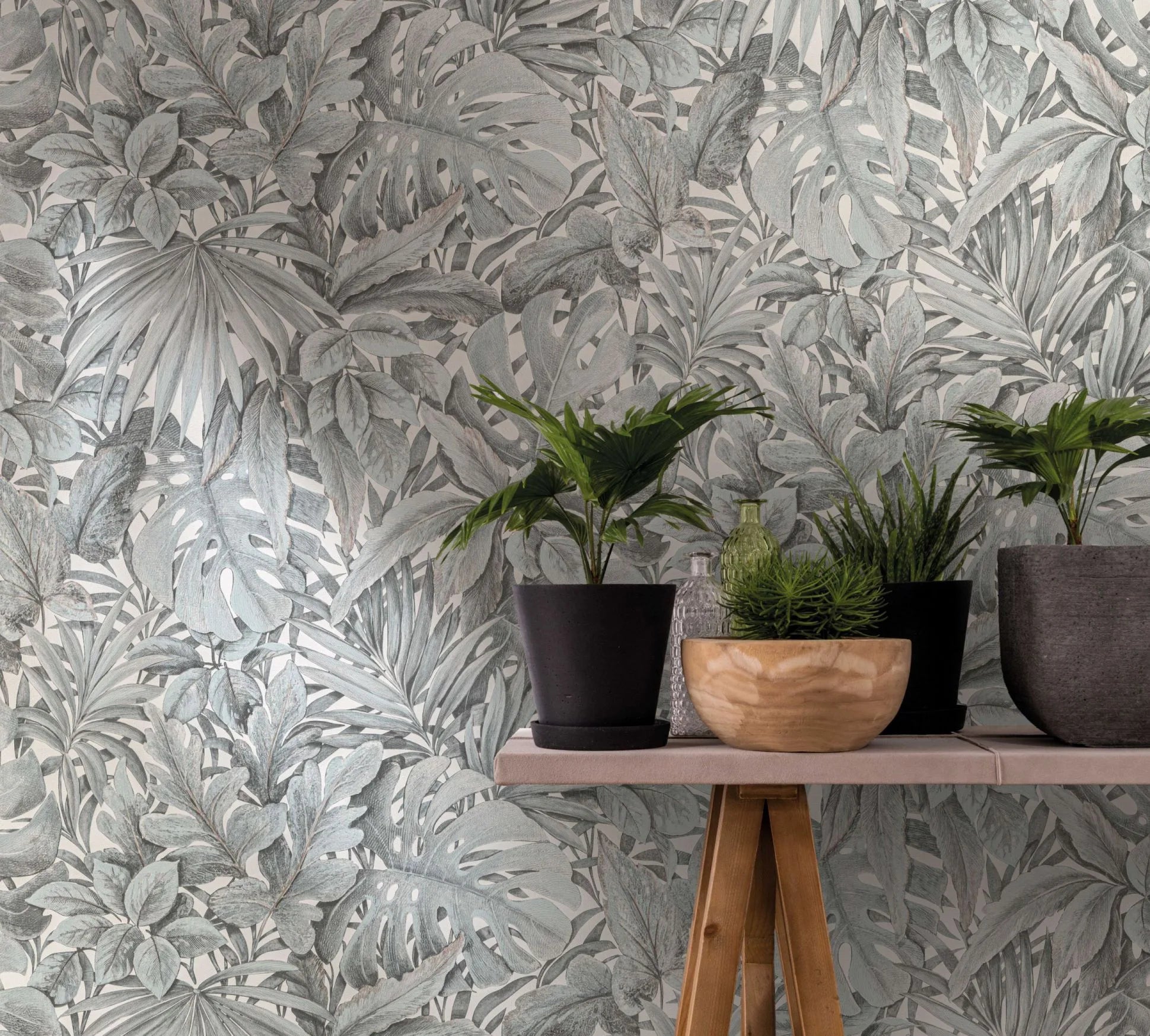 Marburg Botanica 33306 Green and white botanical wallpaper with layered tropical leaves, shown behind a light wood console holding several potted plants, a wooden bowl, and small glass bottles.