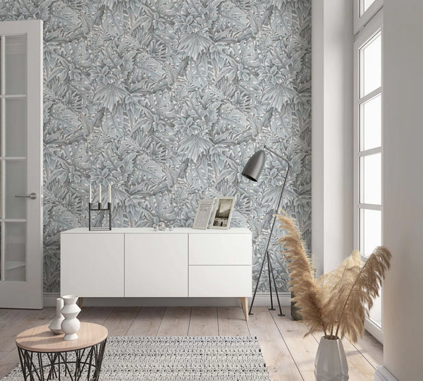 Marburg Botanica 33306 Tropical leaf wallpaper in soft gray tones used as a feature wall behind a white sideboard, with a floor lamp, small round table, and a tall vase with pampas grass beside a window.