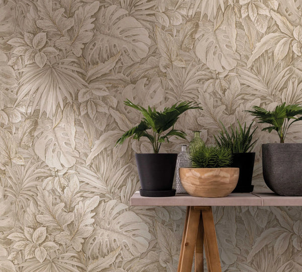 Marburg Botanica 33307 Beige tropical-leaf wallpaper with overlapping foliage. In front, a narrow tabletop on a wooden trestle holds several potted plants, a wooden bowl, and two glass bottles.