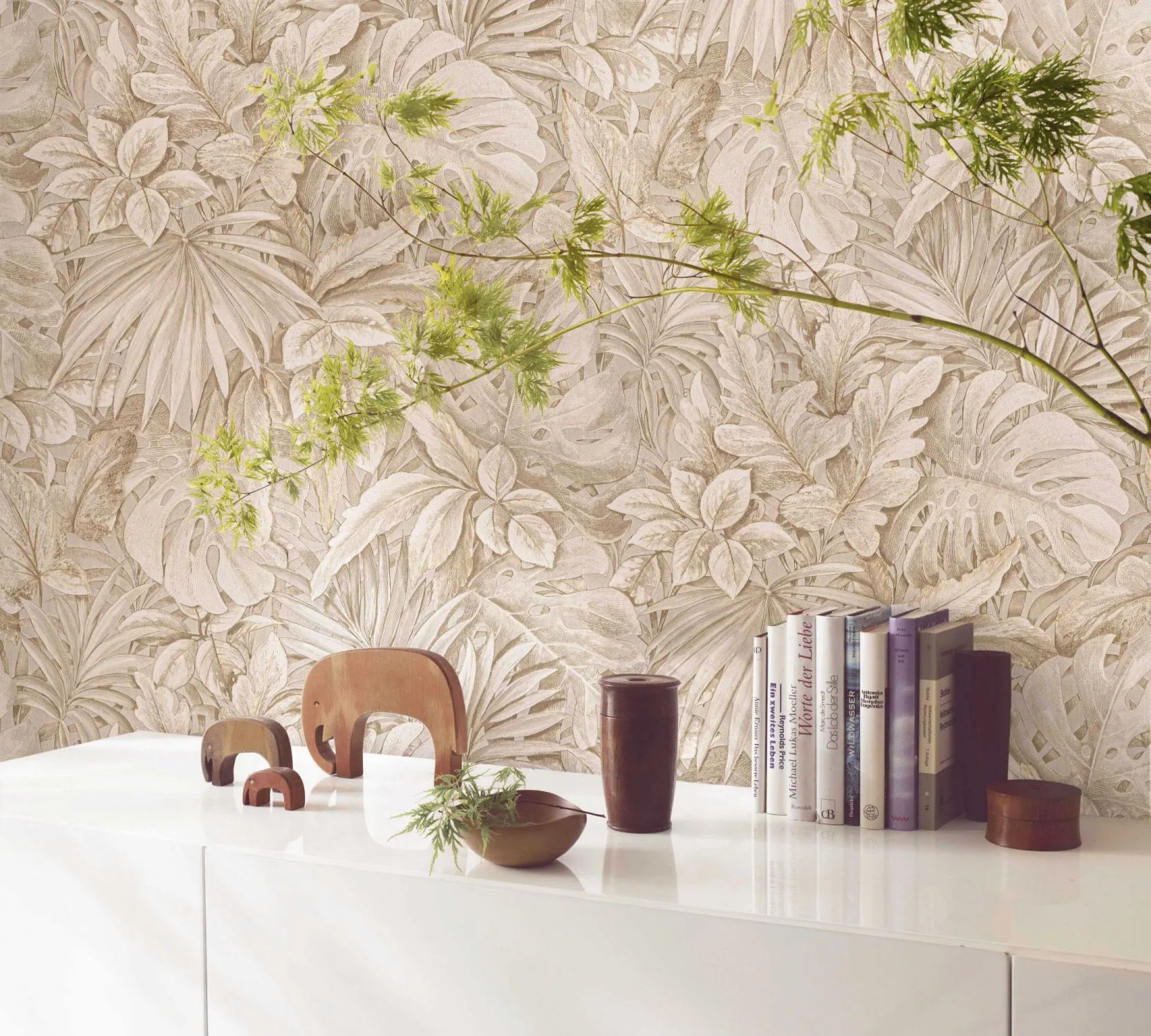 Marburg Botanica 33307 Beige tropical foliage wallpaper behind a white sideboard styled with wooden elephant figurines, books, a brown cup, small bowls, and a leafy green branch.