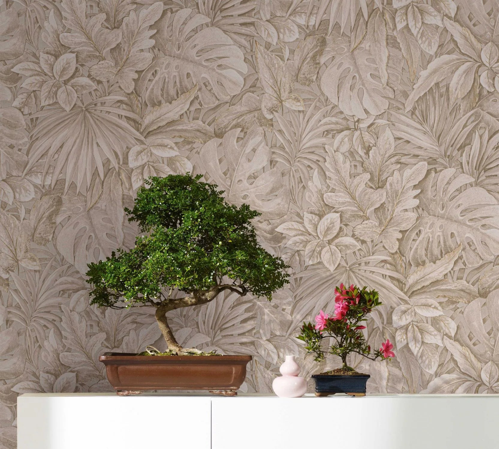 Marburg Botanica 33307 Beige botanical leaf wallpaper forms the backdrop for a scene with a bonsai tree, a smaller flowering bonsai, and a small pink vase on a white cabinet.