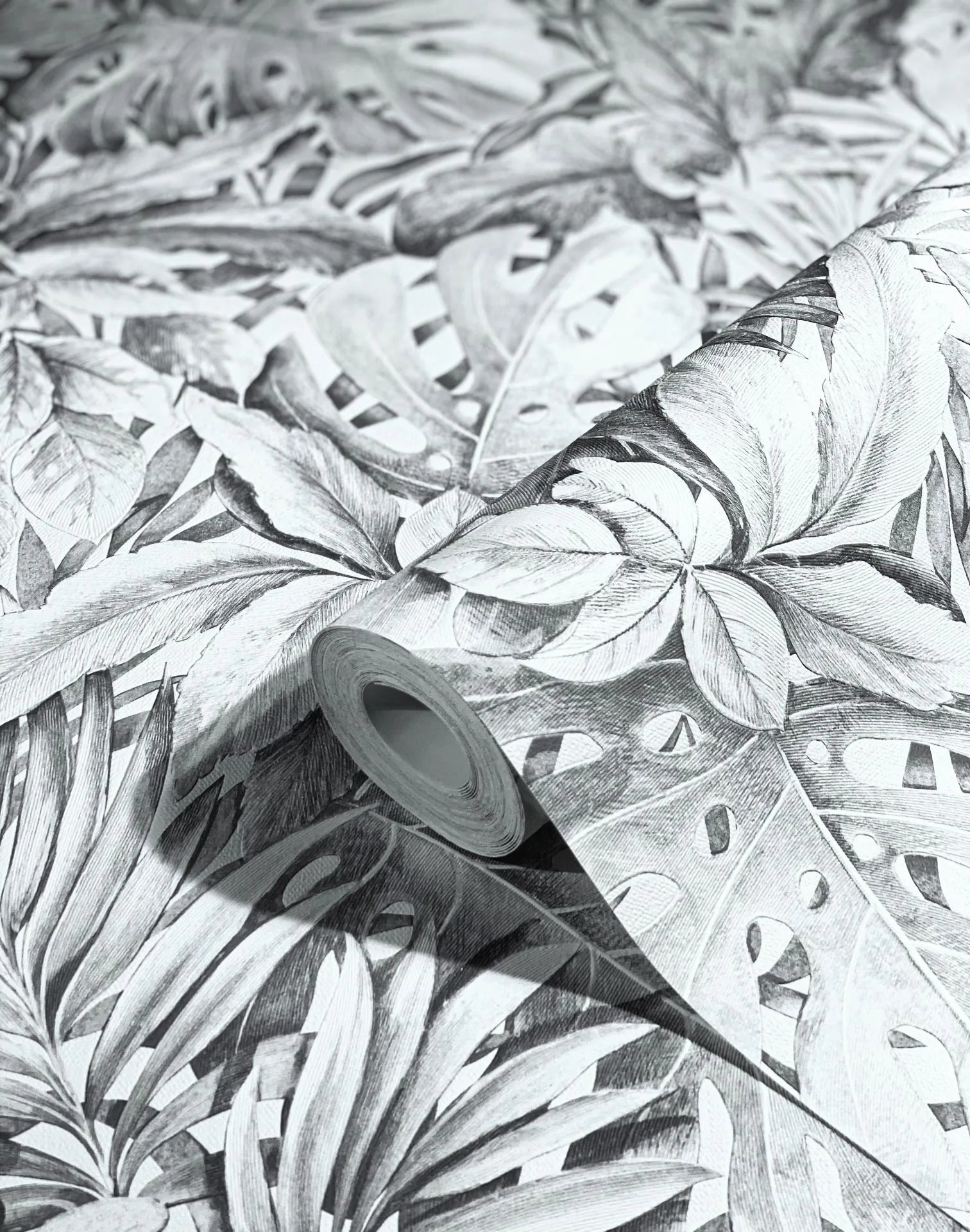 Marburg Botanica 33308 Wallpaper roll resting on matching gray botanical leaf wallpaper, showing a monochrome tropical foliage pattern with large overlapping leaves.