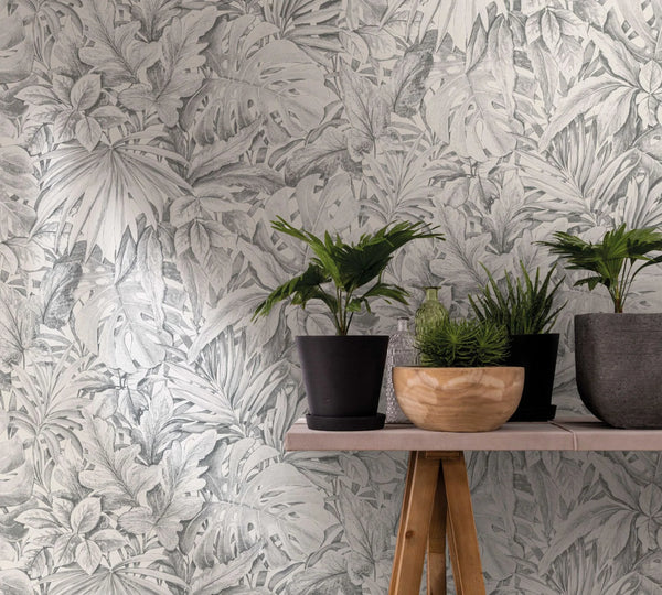 Marburg Botanica 33308 Gray tropical leaf wallpaper behind a small console table styled with potted plants, a wooden bowl, and glass bottles.
