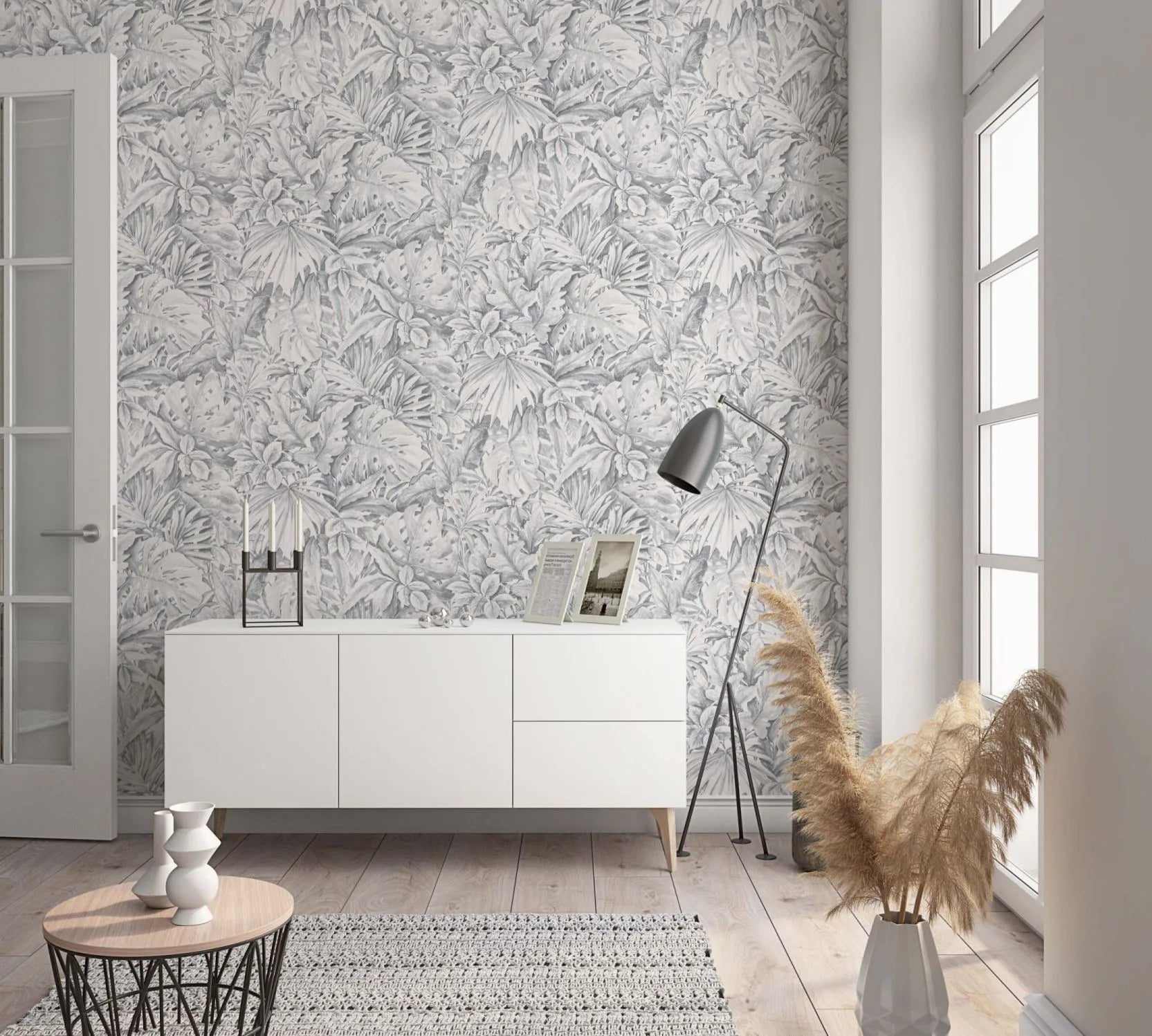 Marburg Botanica 33308 Living room scene featuring gray botanical leaf wallpaper behind a white sideboard with framed photos and a candlestick, a black floor lamp, a small round coffee table, a rug, and a vase of pampas grass by a tall window.
