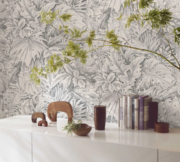 Marburg Botanica 33308 Gray tropical foliage wallpaper with large sketched leaves behind a white cabinet topped with books, wooden elephant figurines, small wooden vessels, and a leafy branch in the foreground.