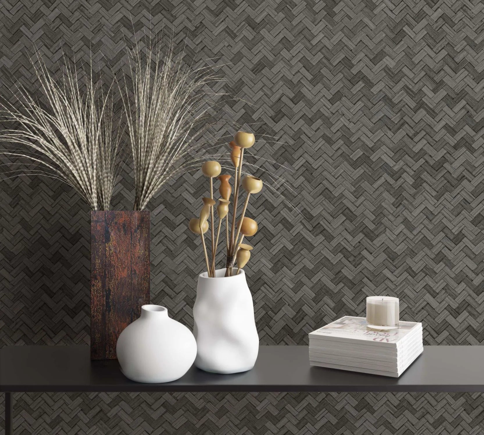 Marburg Botanica 33310 Dark gray woven herringbone wallpaper behind a black console holding a rust-toned tall vase with grasses, two white ceramic vases with dried stems, and a stack of magazines topped with a small candle.