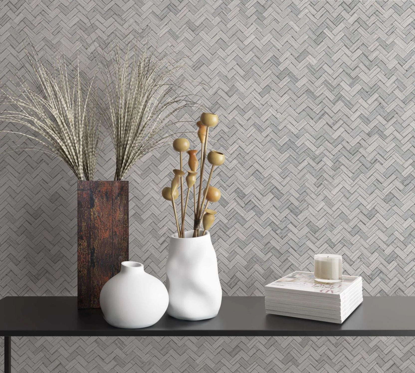 Marburg Botanica 33311 Beige and gray herringbone-pattern wallpaper behind a black shelf holding decorative vases with dried stems, a candle, and a stacked magazine set.