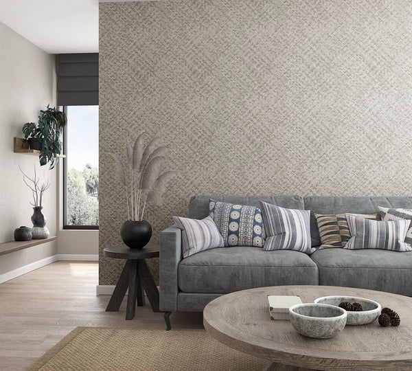 Marburg Botanica 33311 Beige and gray woven geometric wallpaper on a feature wall behind a gray sofa with patterned cushions, a round wood coffee table with small bowls and pinecones, and a side table with a black vase of dried grass, with a window and floating shelf visible.