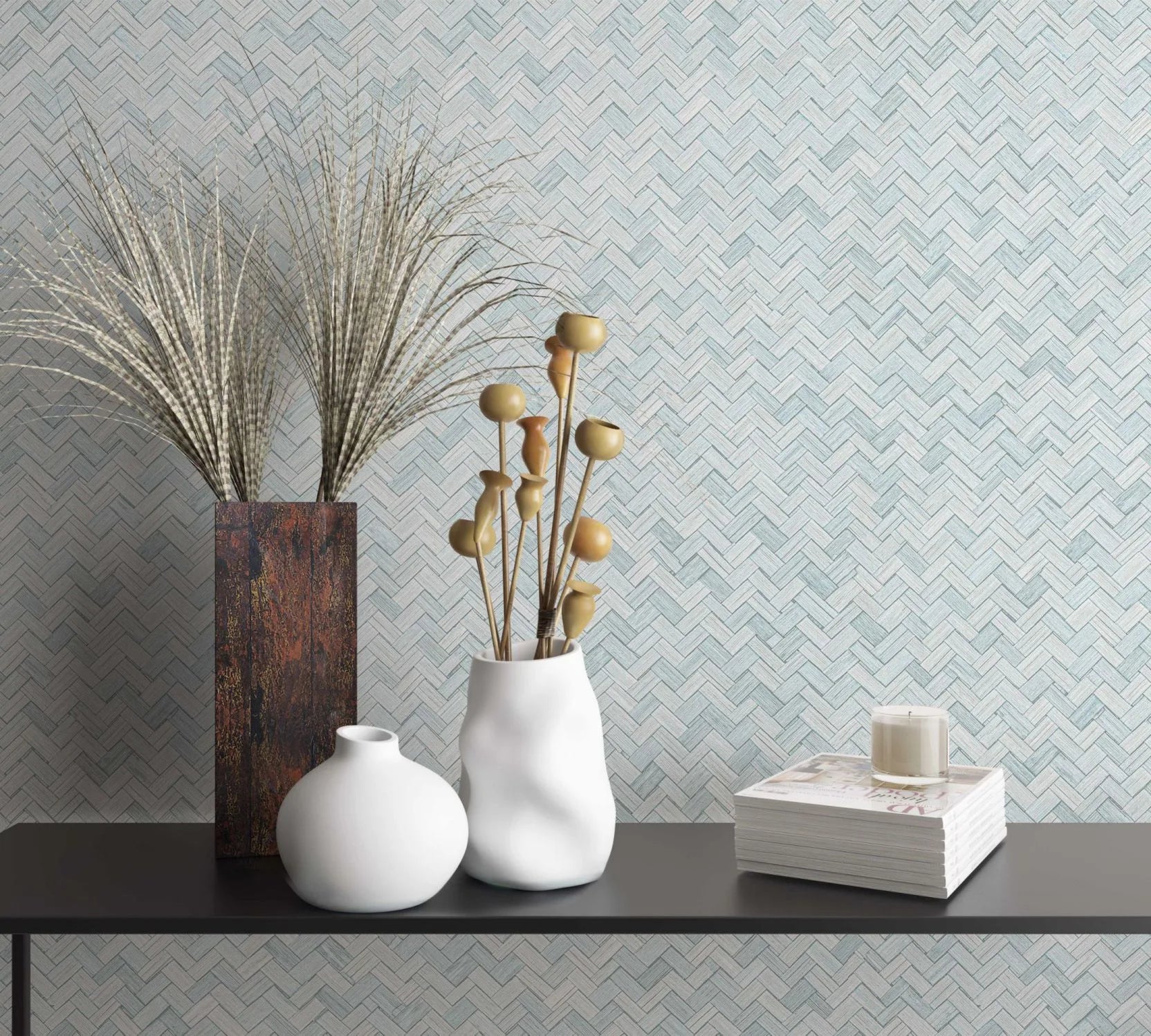 Marburg Botanica 33312 Pale green herringbone wallpaper behind a black console with two white vases holding dried stems, a tall rectangular vase with grasses, and a stack of magazines topped with a candle.