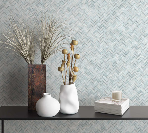 Marburg Botanica 33312 Pale green herringbone wallpaper behind a black console with two white vases holding dried stems, a tall rectangular vase with grasses, and a stack of magazines topped with a candle.