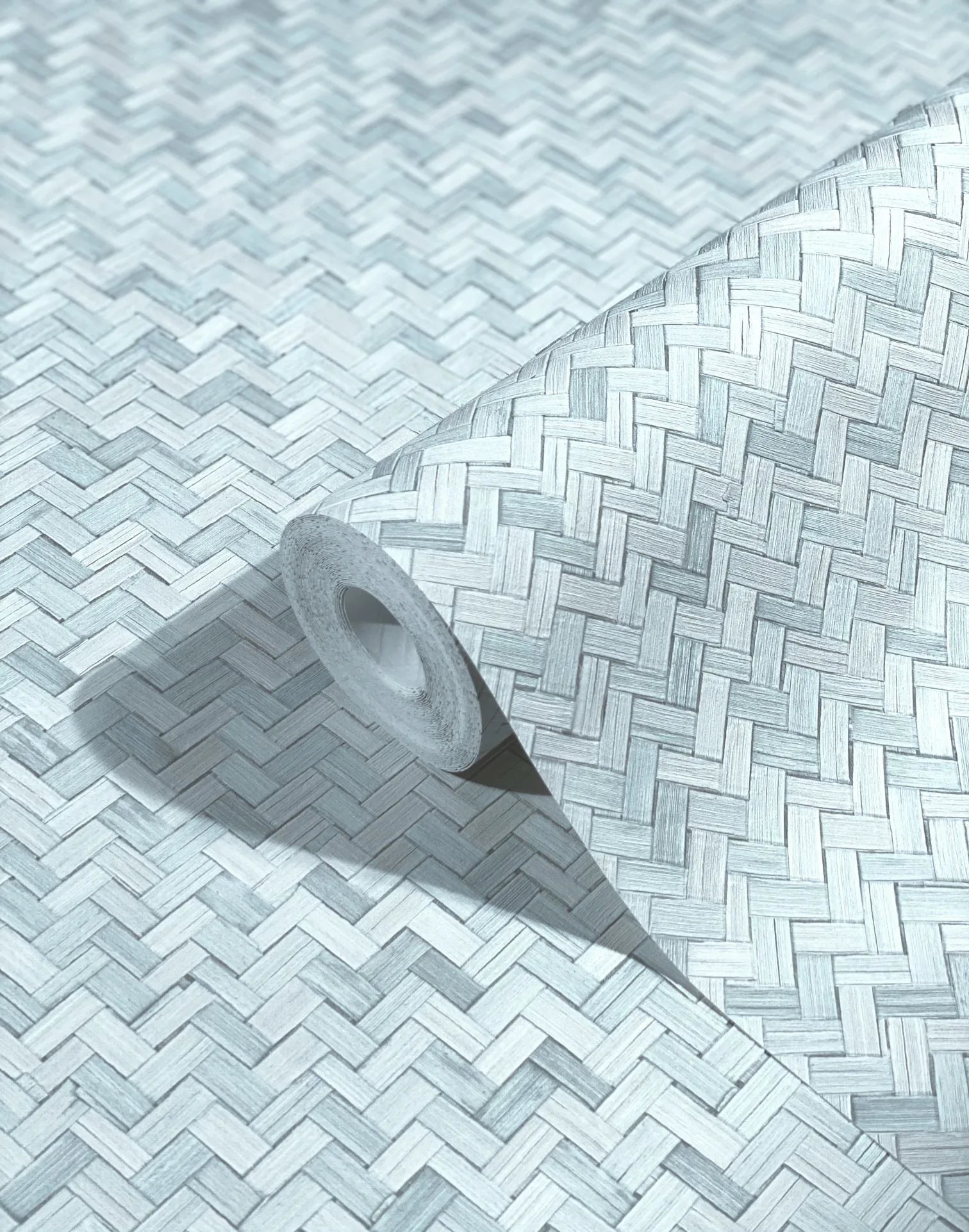 Marburg Botanica 33312 Angled wallpaper roll on a matching surface showing an interlaced herringbone weave pattern in light blue‑gray tones.