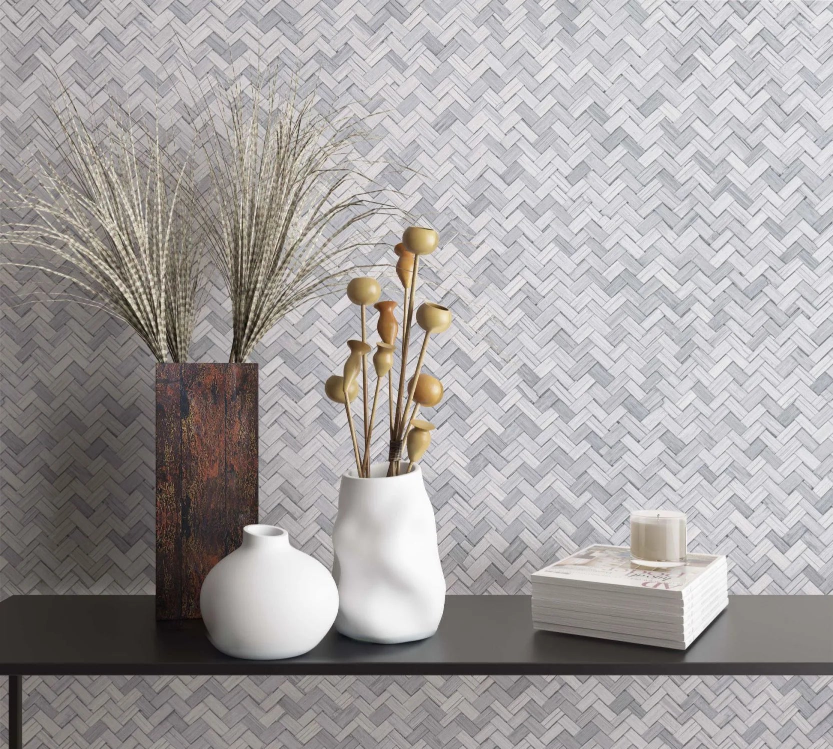 Marburg Botanica 33314 Gray herringbone wallpaper behind a black console holding a tall rectangular vase with grasses, two white vases—one with dried seed pods—and a stack of magazines with a small candle.