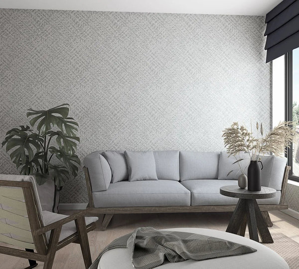 Marburg Botanica 33314 Wide view of a living room featuring light gray crosshatch wallpaper behind a gray sofa, with a round wooden side table holding a black vase of dried grasses, an armchair, a potted plant, a window with a dark shade, and an ottoman with a throw.