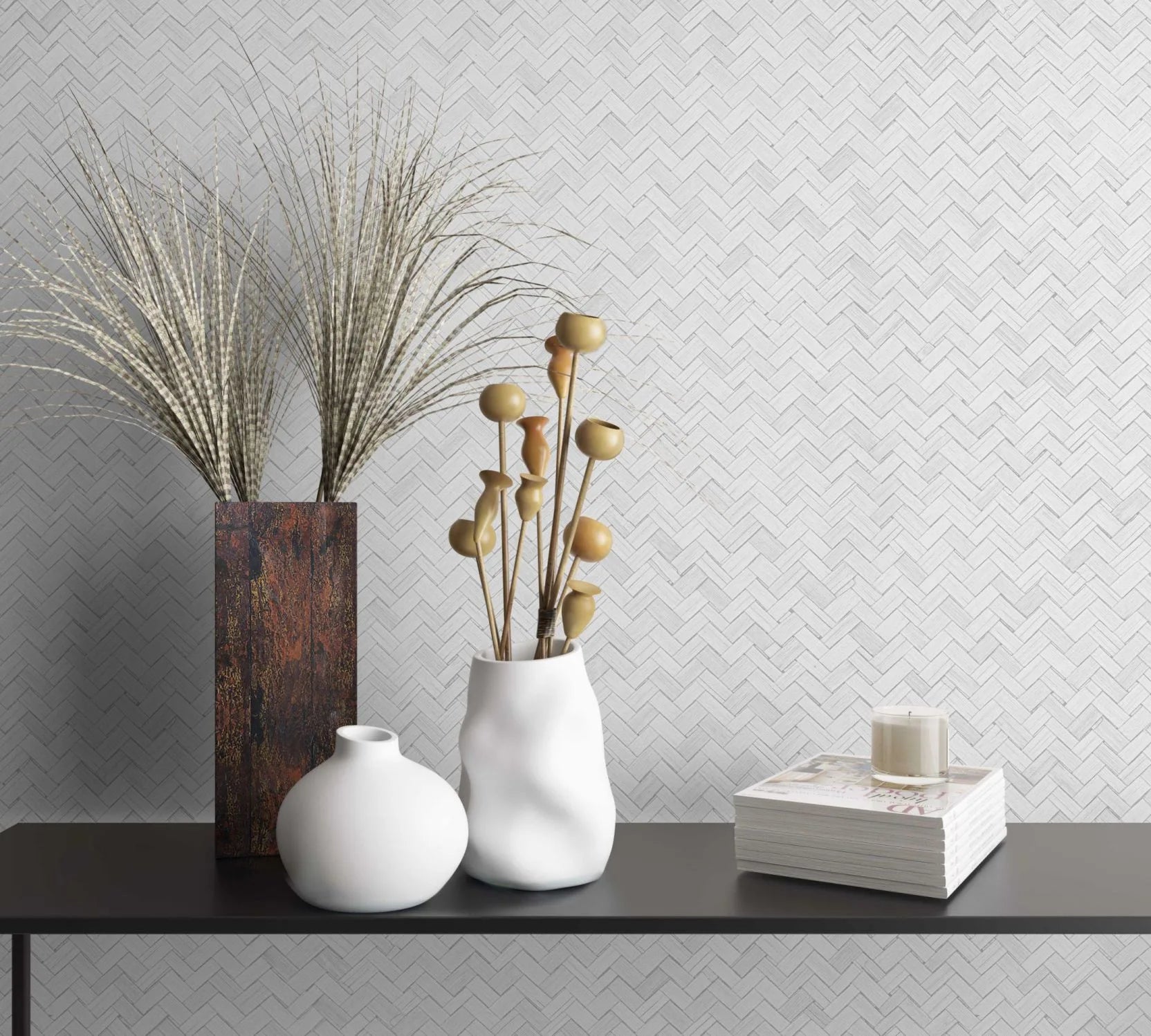Marburg Botanica 33315 White herringbone wallpaper behind a black console holding two white vases, a tall rectangular rust-toned vase with grasses, a stack of magazines, and a candle.
