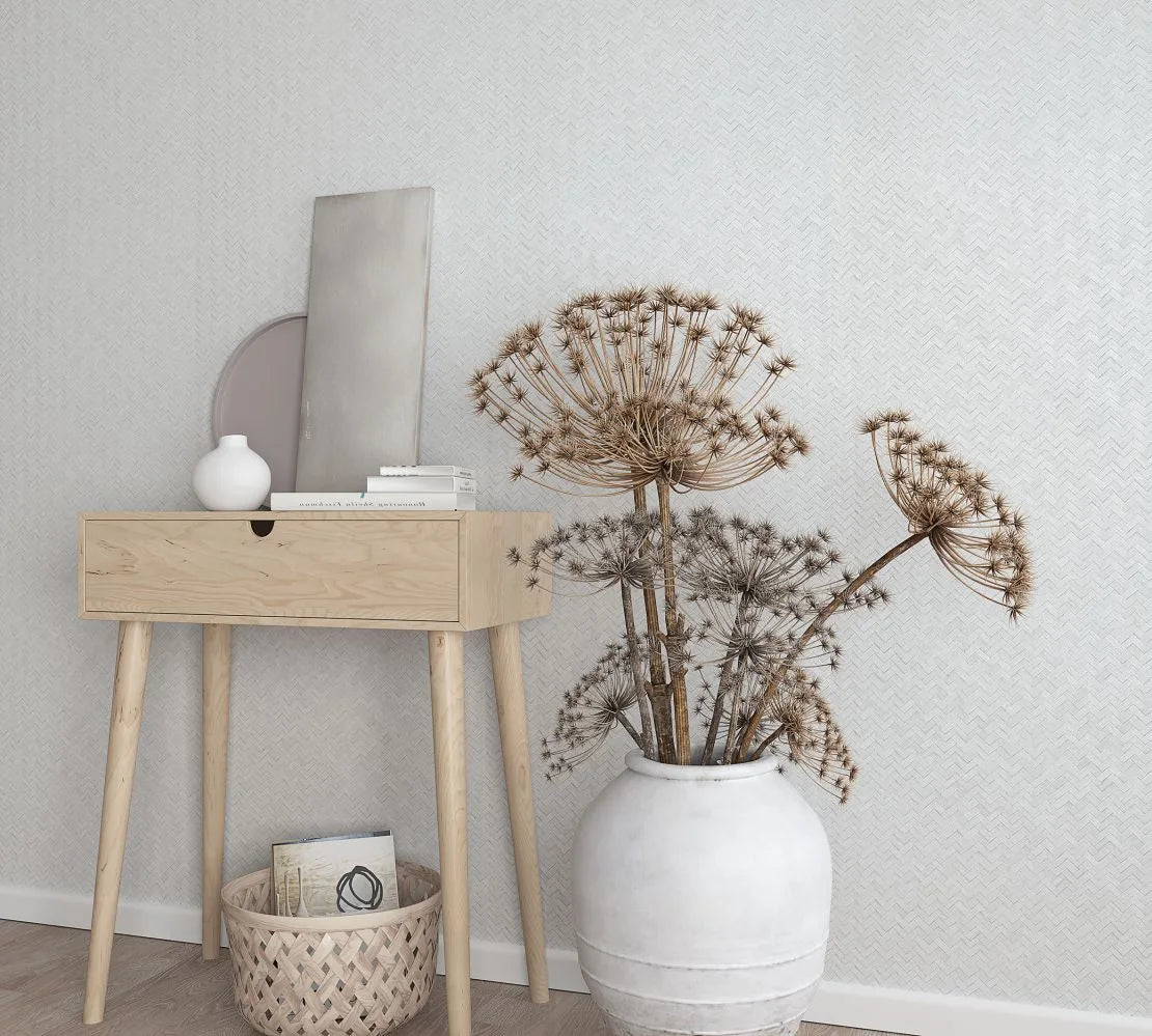 Marburg Botanica 33315 Light herringbone wallpaper behind a small wooden console with decor, a woven basket on the floor, and a large white ceramic pot holding dried flower stems.