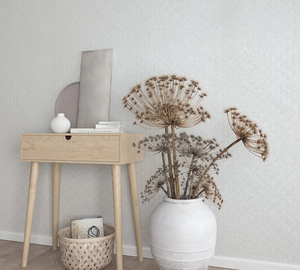 Marburg Botanica 33315 Light herringbone wallpaper behind a small wooden console with decor, a woven basket on the floor, and a large white ceramic pot holding dried flower stems.