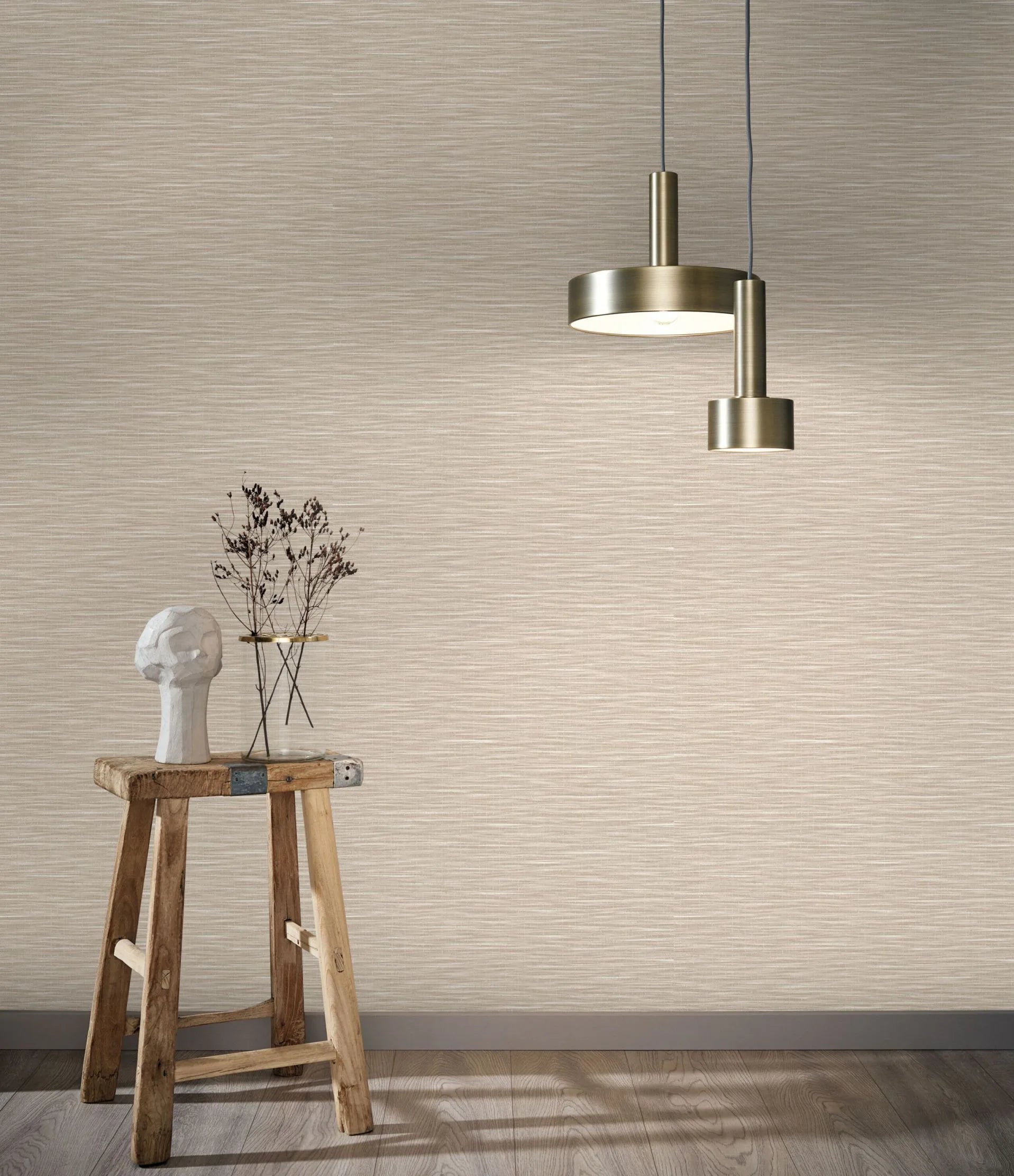 Marburg Botanica 33316 Room scene showing beige textured wallpaper with a horizontal strie pattern, two brass pendant lights, and a rustic wooden stool holding a sculptural head and a small glass vessel with dried stems; wood floor and baseboard visible.