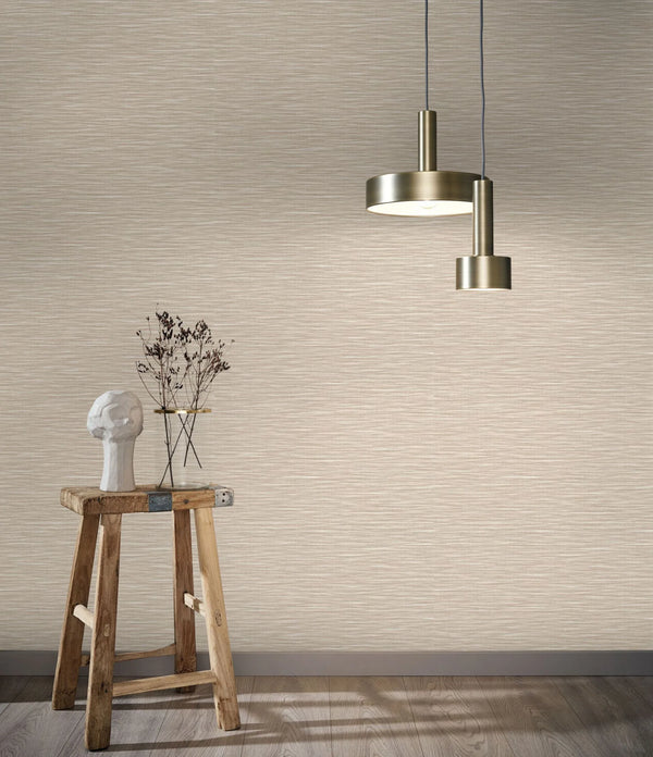 Marburg Botanica 33316 Room scene showing beige textured wallpaper with a horizontal strie pattern, two brass pendant lights, and a rustic wooden stool holding a sculptural head and a small glass vessel with dried stems; wood floor and baseboard visible.