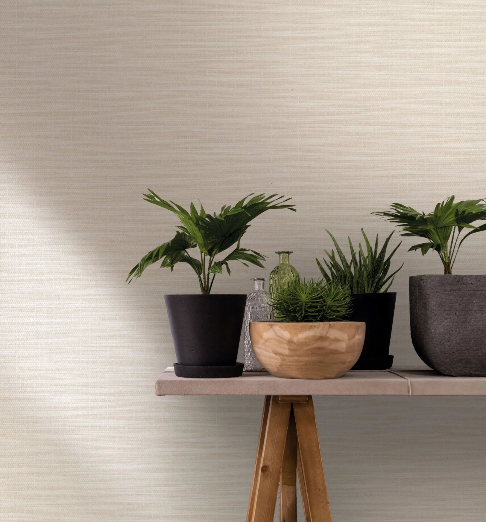 Marburg Botanica 33318 Beige and white wallpaper with subtle horizontal striations. In front, a trestle table holds potted plants, a wooden bowl, and clear and green glass bottles.