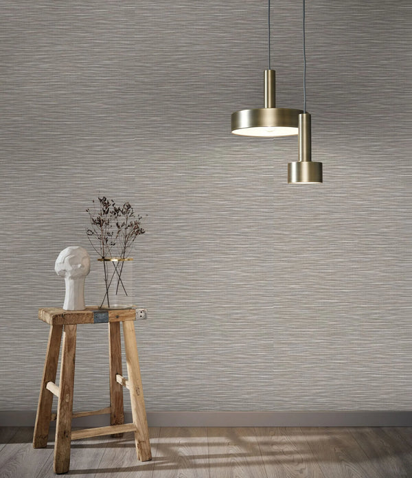 Marburg Botanica 33319 Neutral brown‑grey wallpaper with fine horizontal strie texture. Two brushed brass pendant lights hang above a rustic wooden stool holding a white bust and a glass jar with dried stems on a wood floor.