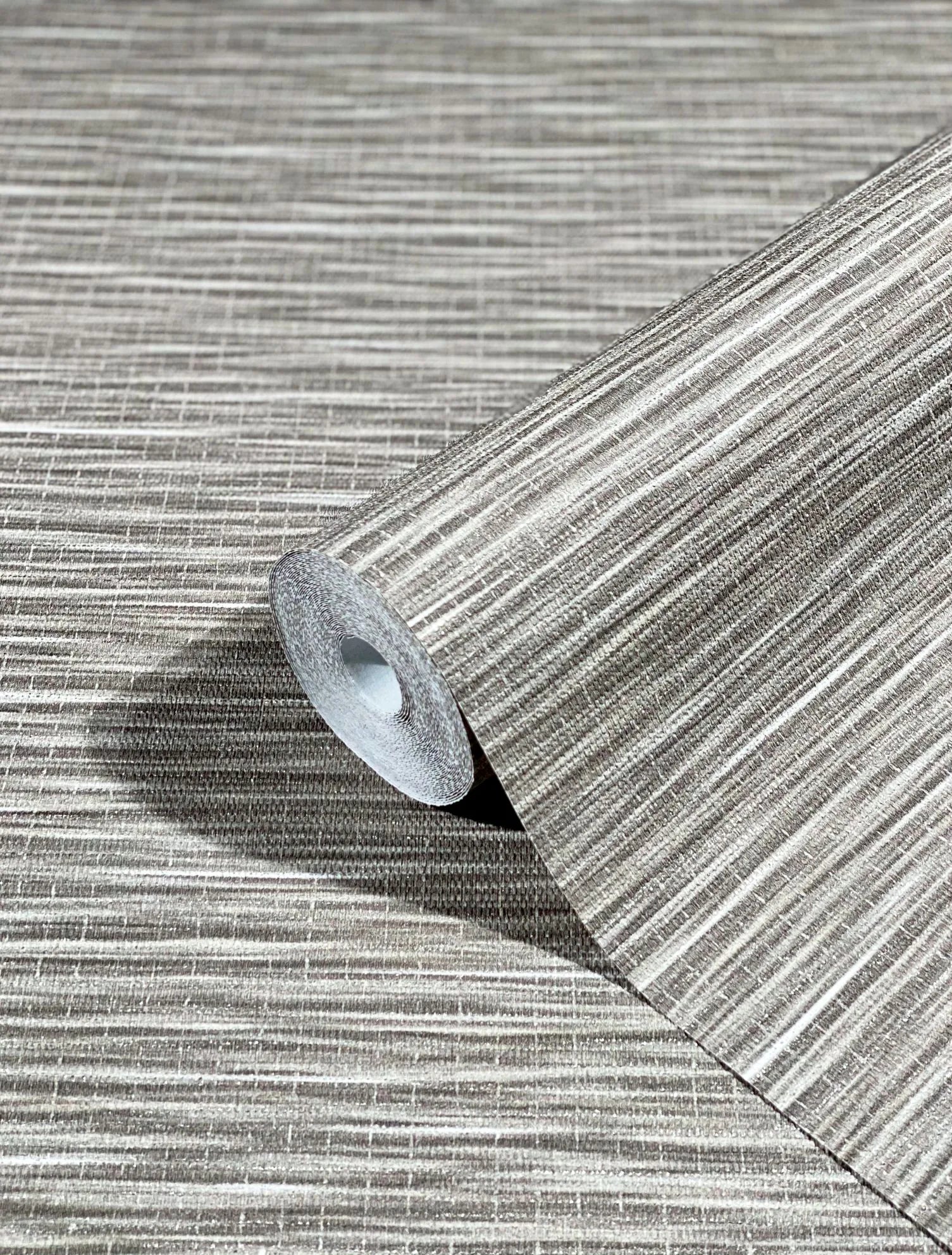 Marburg Botanica 33319 Close-up of a wallpaper roll resting on a matching unrolled sheet, showing a neutral brown linear striated pattern with a subtle crosshatch texture.