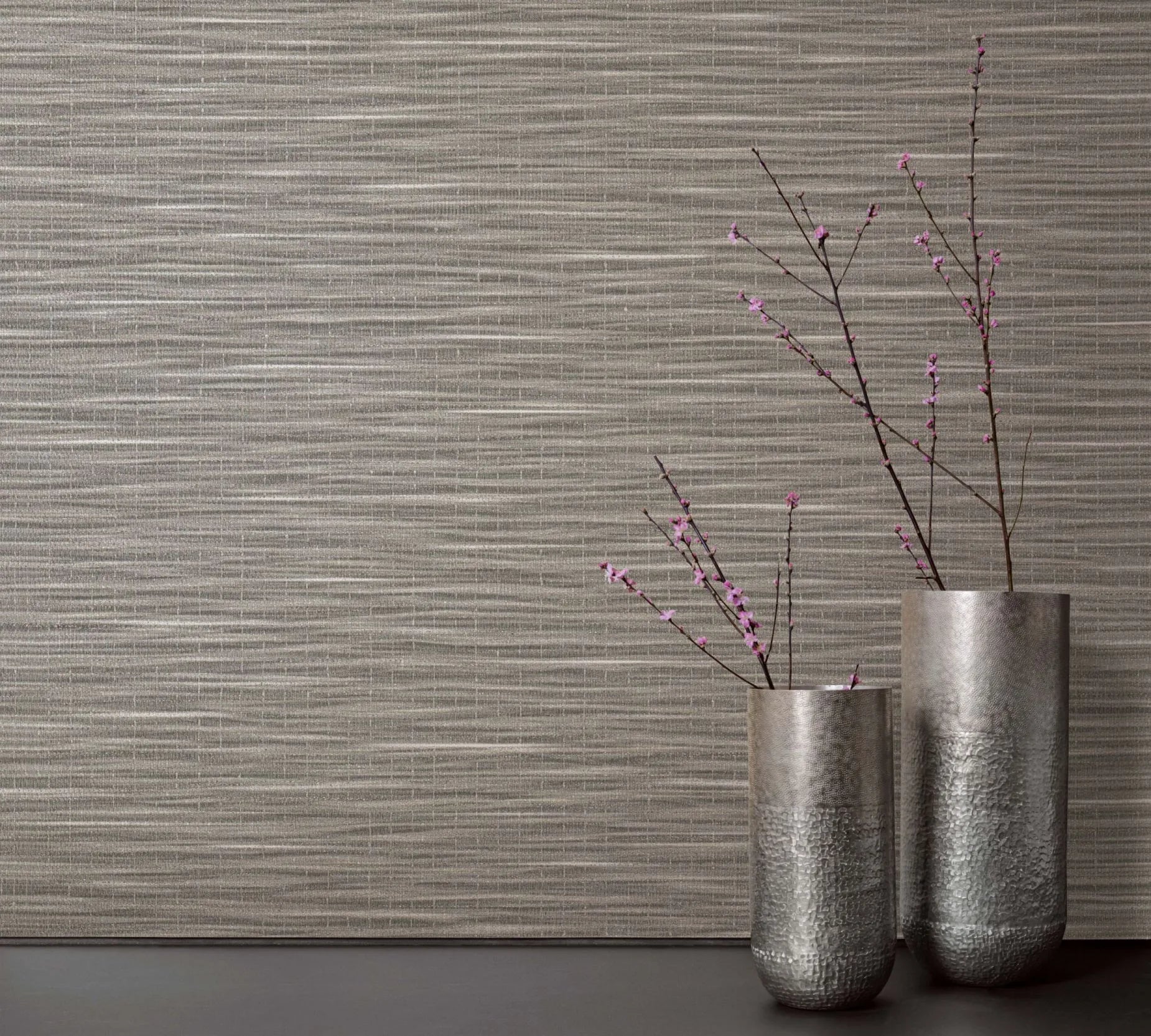 Marburg Botanica 33319 Textured brown wallpaper with horizontal striations, styled with two tall silver vases holding branches with small pink blossoms on a dark surface.