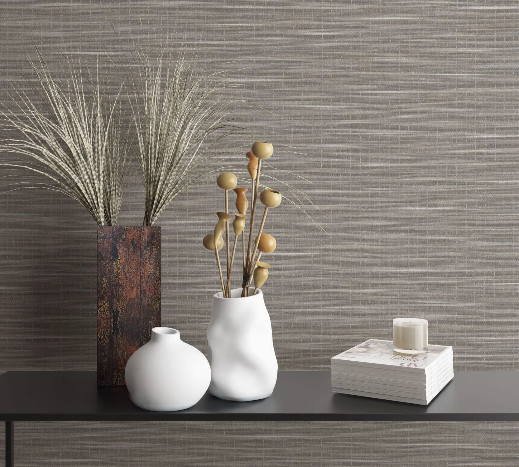 Marburg Botanica 33319 Horizontal striated brown‑taupe wallpaper behind a black shelf holding a rust‑colored rectangular vase with grasses, two white ceramic vases with dried stems, and a stack of magazines topped with a small candle.