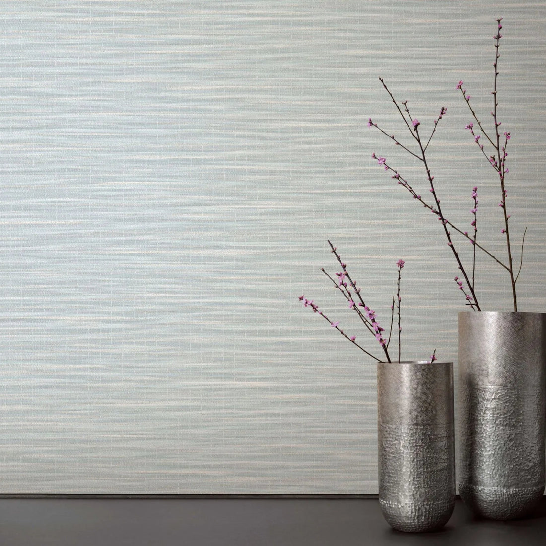 Marburg Botanica 33321 Light green and white horizontally striated wallpaper behind two tall metal vases with pink flowering branches on a dark surface.