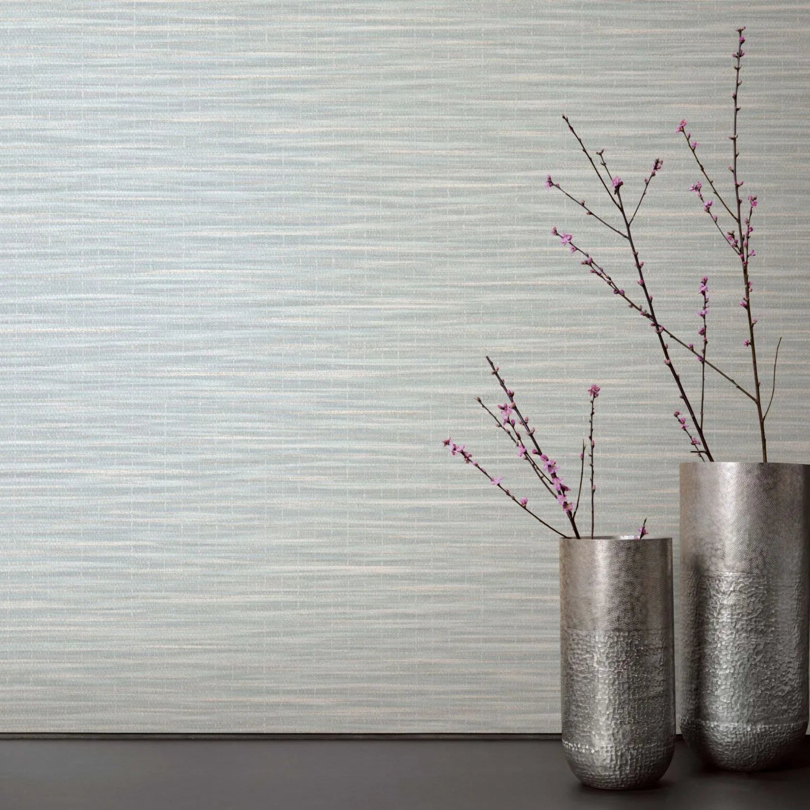 Marburg Botanica 33321 Light green and white horizontally striated wallpaper behind two tall metal vases with pink flowering branches on a dark surface.