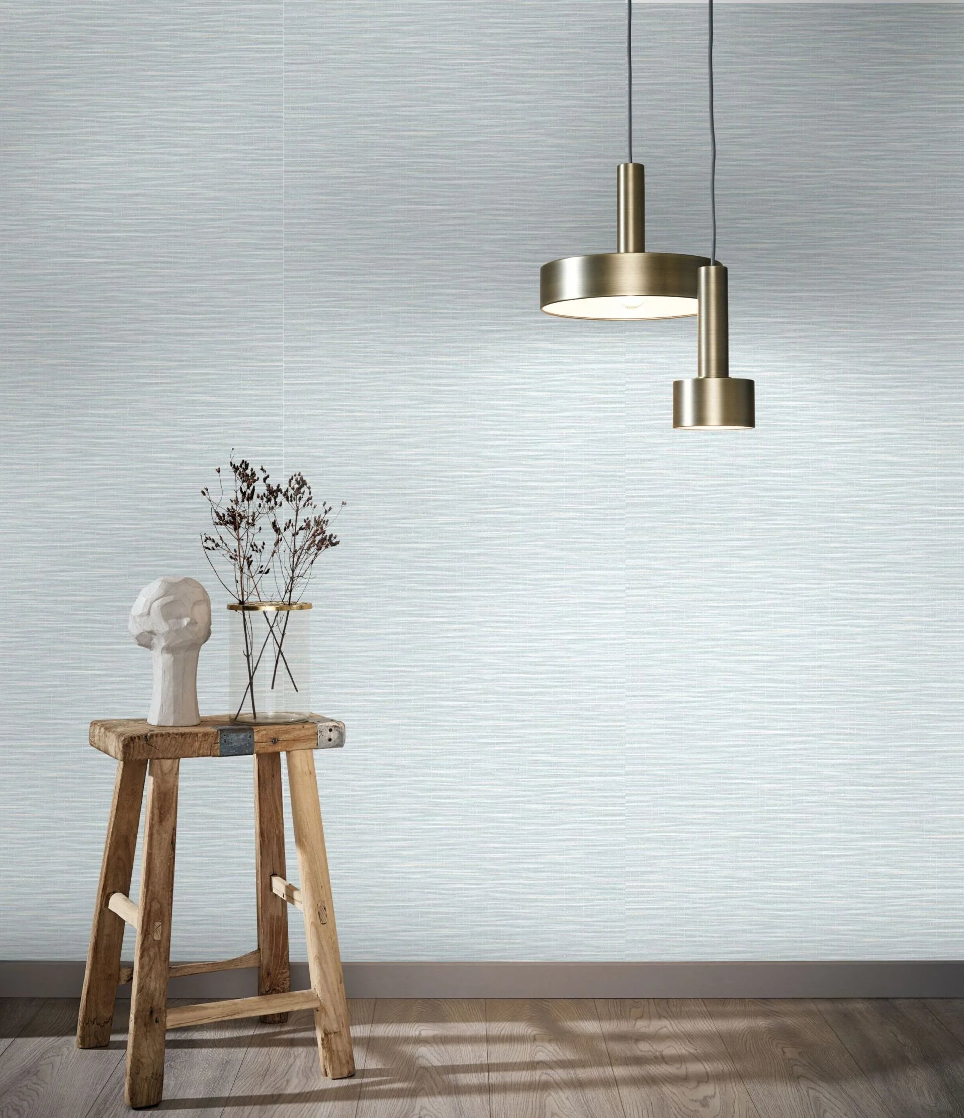 Marburg Botanica 33321 Pale neutral wallpaper with fine horizontal striations shown in a styled scene with a brass pendant light, a rustic wooden stool holding a small sculpture and a glass vase with branches, and a wood floor with baseboard.
