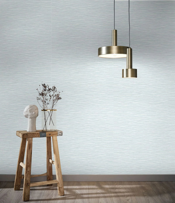 Marburg Botanica 33321 Pale neutral wallpaper with fine horizontal striations shown in a styled scene with a brass pendant light, a rustic wooden stool holding a small sculpture and a glass vase with branches, and a wood floor with baseboard.