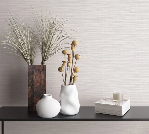 Marburg Botanica 33322 Light gray and white wallpaper with fine horizontal striations behind a black console holding a tall rectangular vase with grasses, two white ceramic vases with dried stems, and a stack of magazines topped with a small candle.