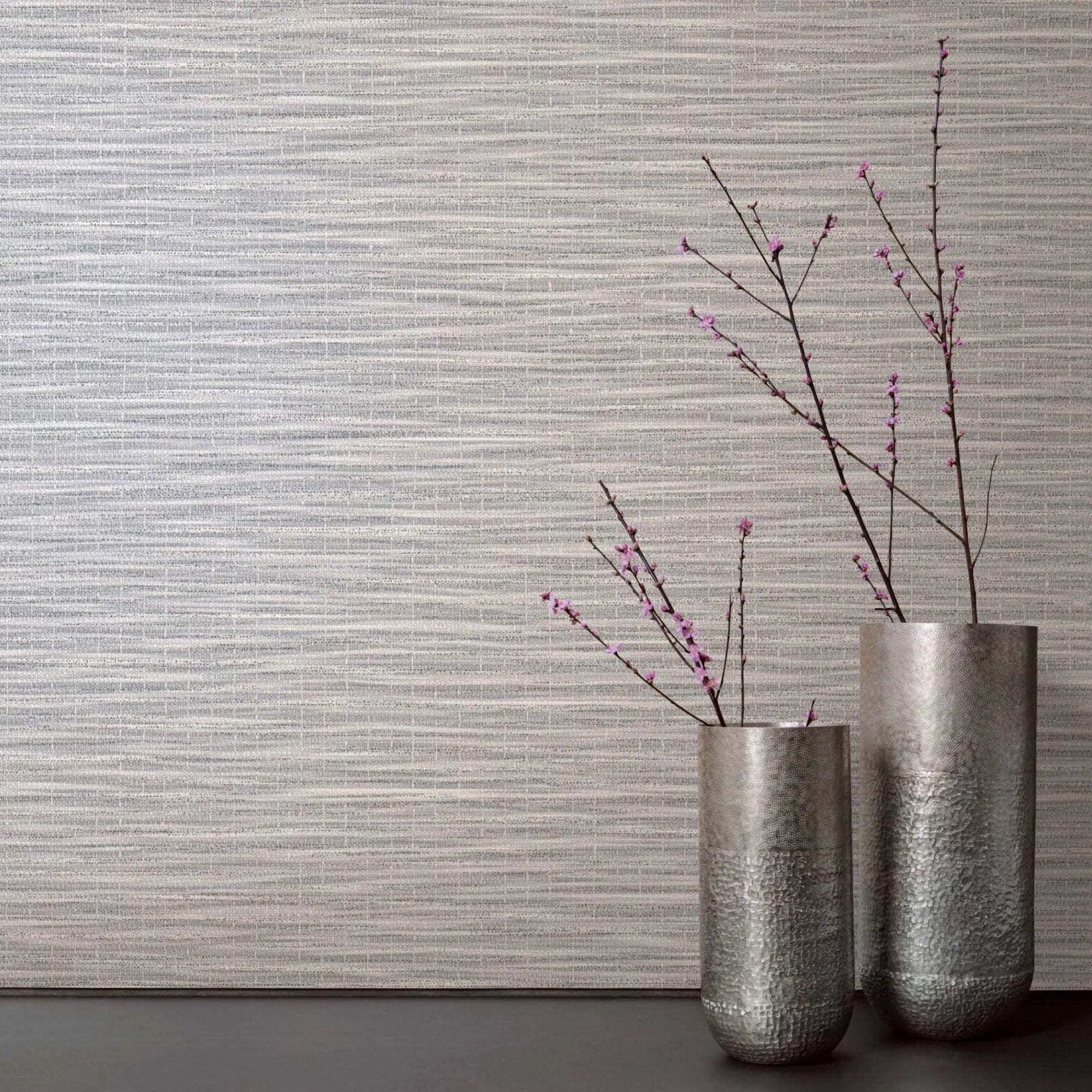 Marburg Botanica 33323 Gray horizontally textured wallpaper shown behind two tall silver vases on a dark surface, with bare branches bearing small pink blossoms.