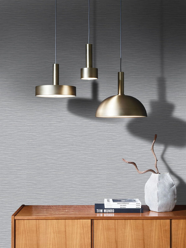 Marburg Botanica 33323 Gray wallpaper with fine horizontal striations behind three brass pendant lights and a wood sideboard with two books and a textured vase holding a branch.