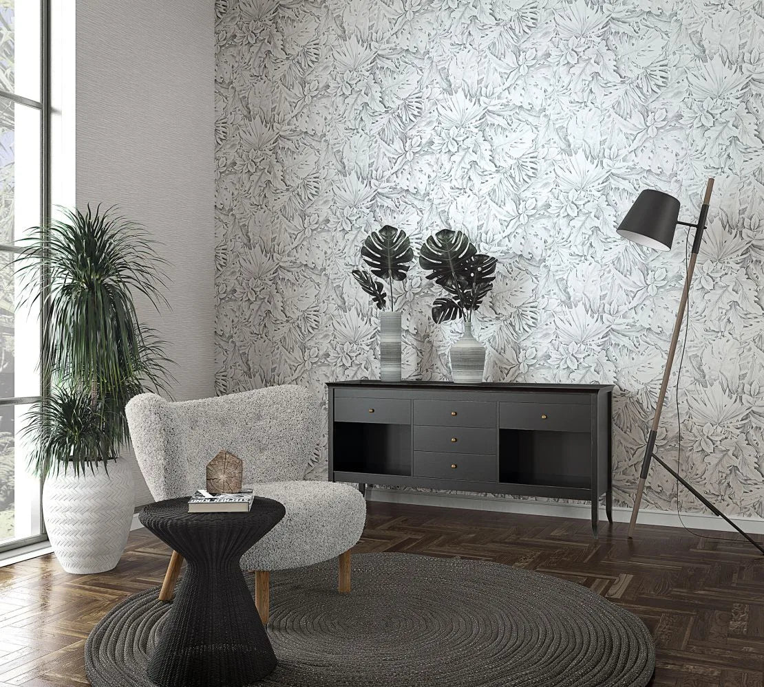 Marburg Botanica 33323 Gray botanical leaf wallpaper on a feature wall behind a dark sideboard with two vases of large leaves, a textured armchair with a black side table, a tall floor lamp, a potted plant by the window, and a round rug on wood flooring.