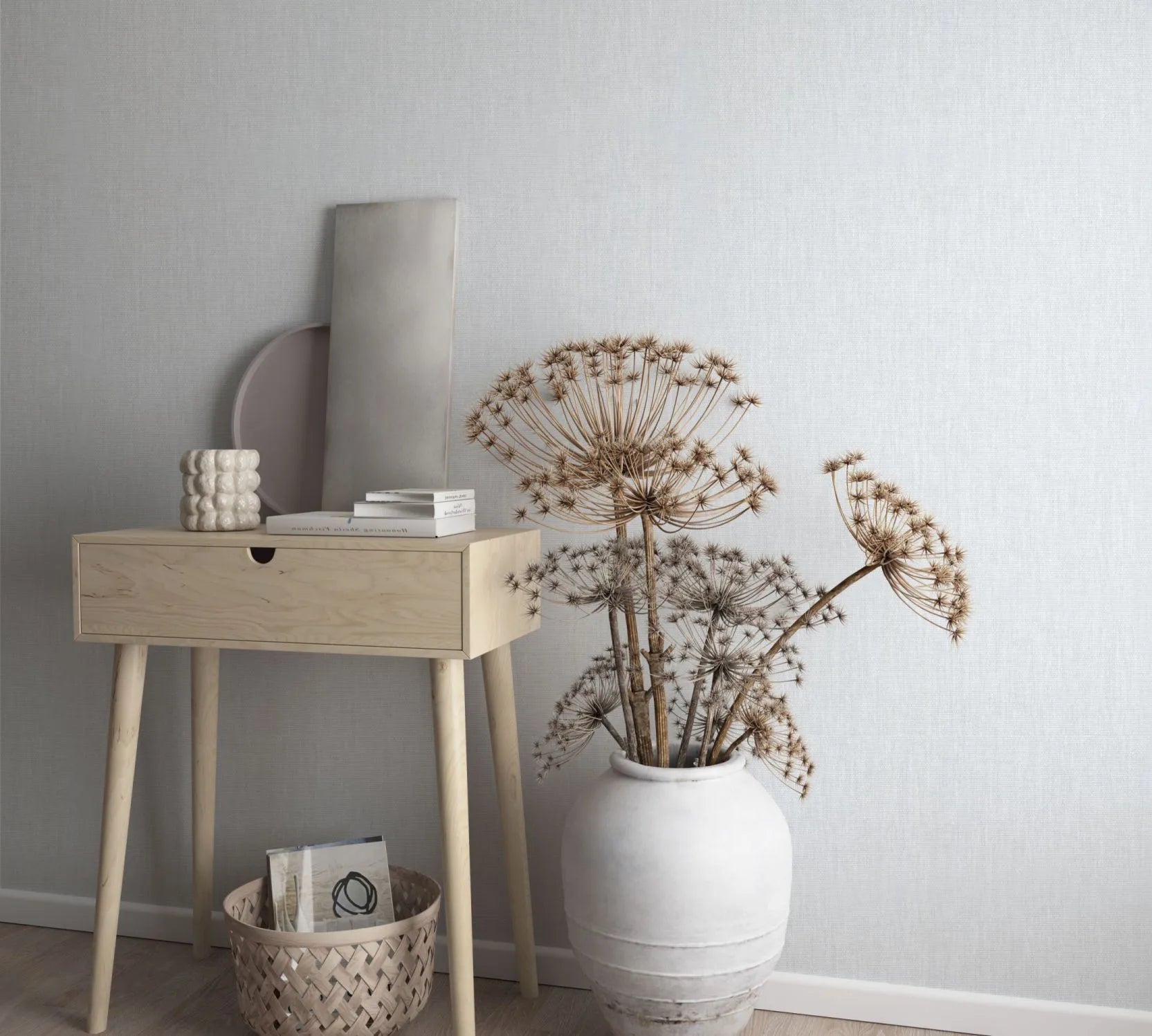 Marburg Botanica 33324 Light gray textured wallpaper behind a light wood console table with books and decor, a floor basket, and a large white vase holding dried stems.