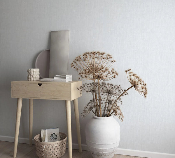 Marburg Botanica 33324 Light gray textured wallpaper behind a light wood console table with books and decor, a floor basket, and a large white vase holding dried stems.