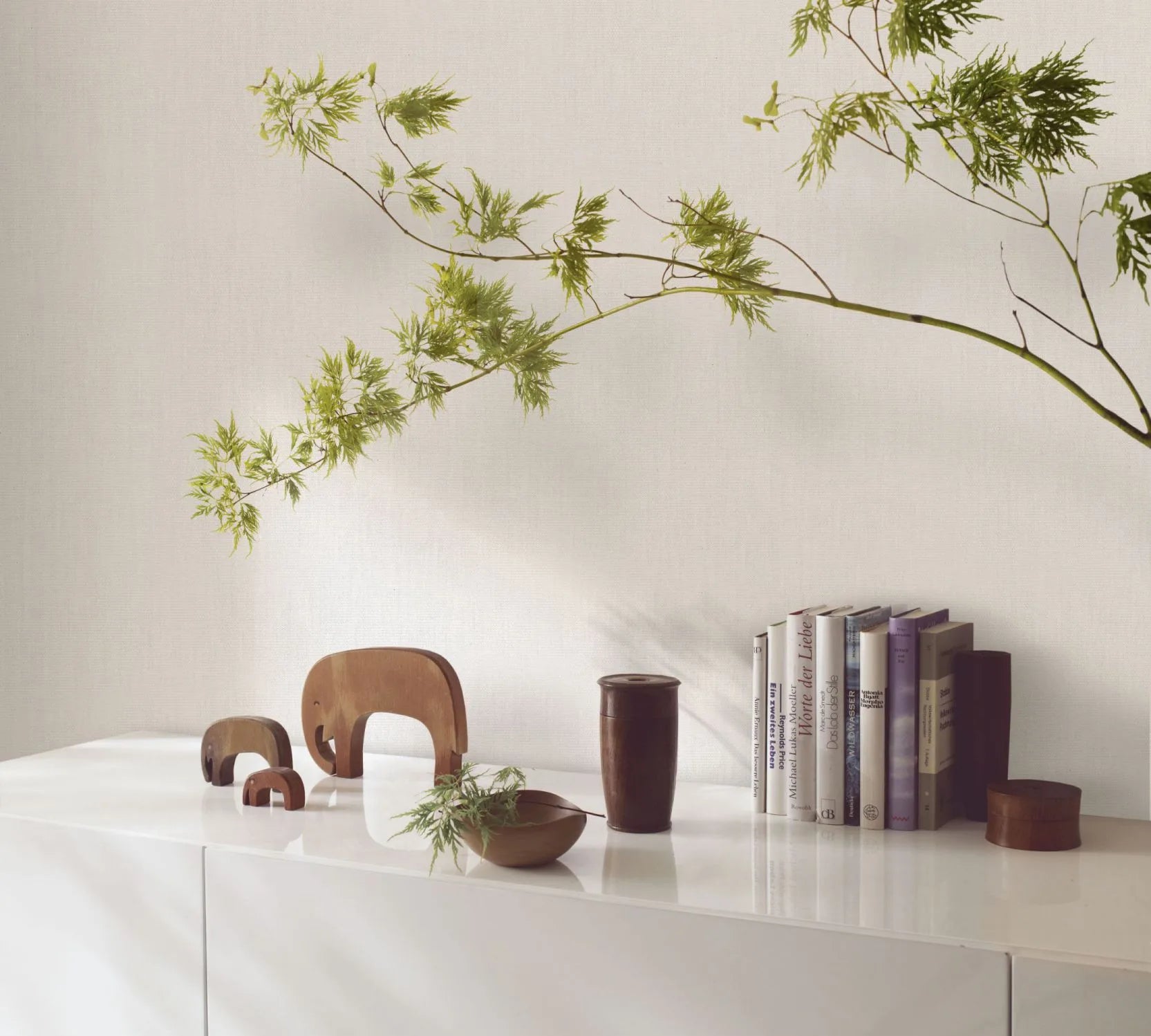 Marburg Botanica 33325 Light gray textured wallpaper with a fine linen look behind a white cabinet styled with wooden animal figures, books, a wooden bowl, and a leafy branch.