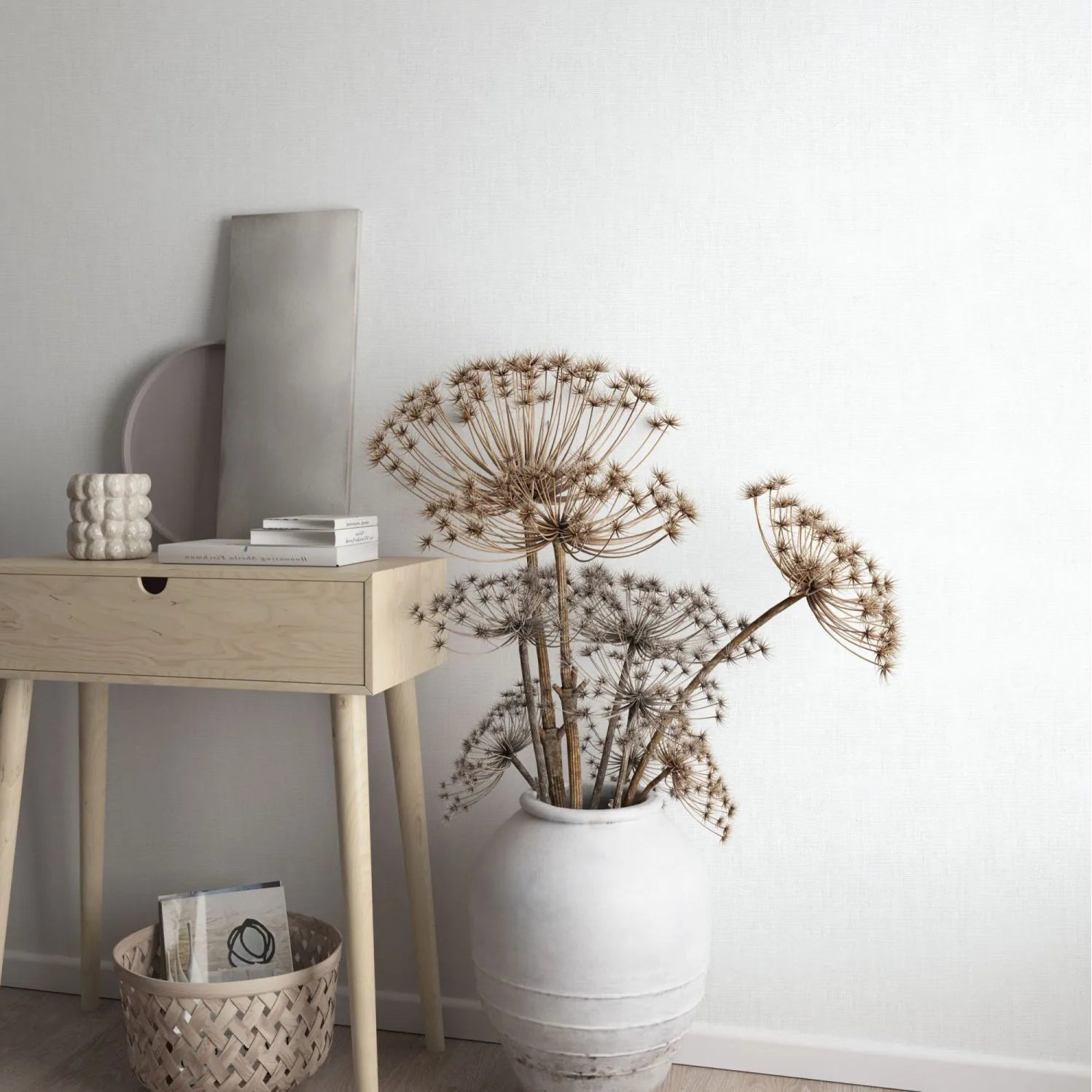 Marburg Botanica 33325 Light gray woven-texture wallpaper behind a light wood console table with books and a sculptural ceramic object, a woven basket on the floor, and a large ceramic vase holding dried seed heads.