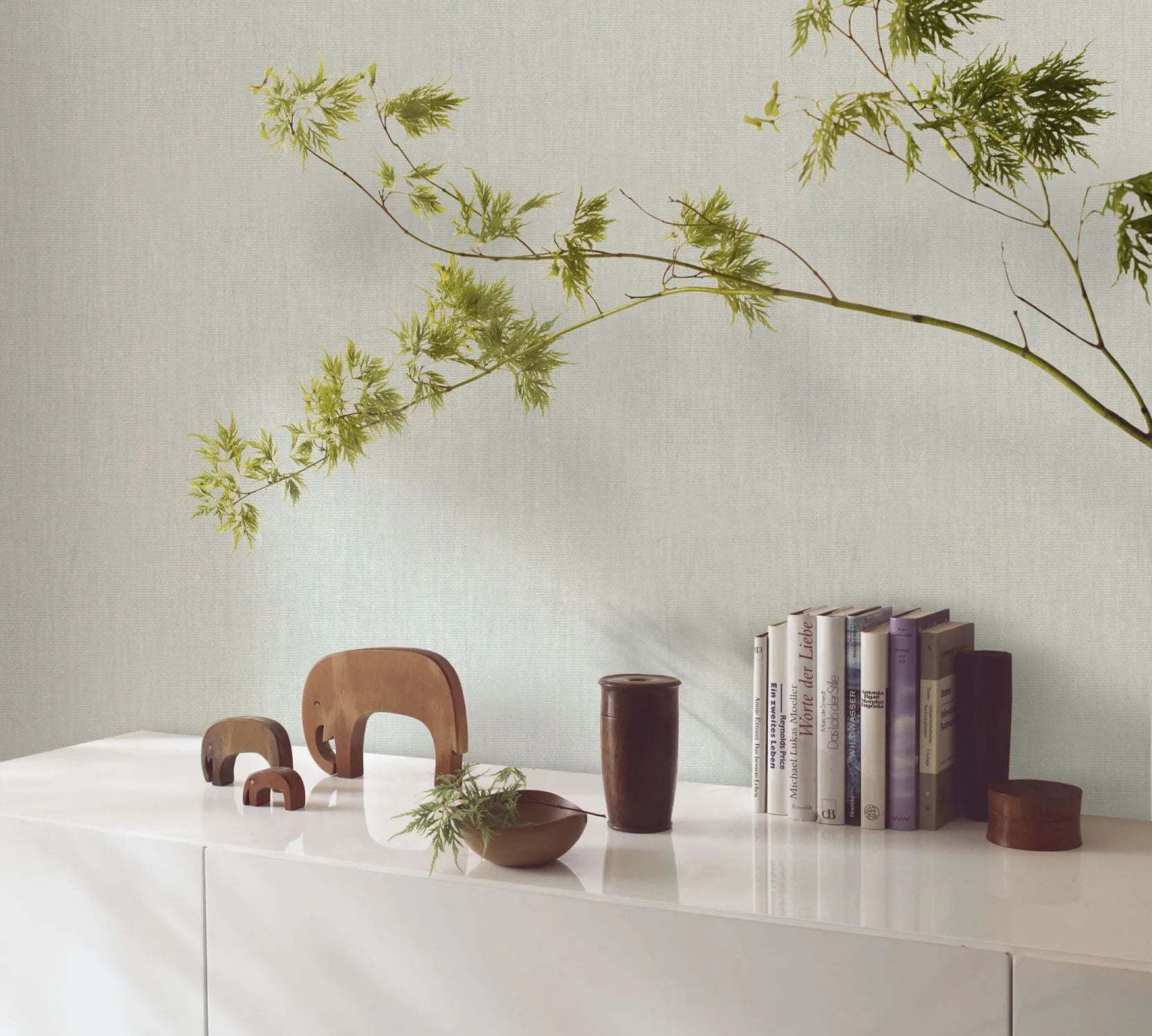 Marburg Botanica 33326 Light green textured wallpaper behind a white sideboard with books, wooden elephant figurines, a wooden cup and bowl, a small canister, and a leafy branch extending across the wall.