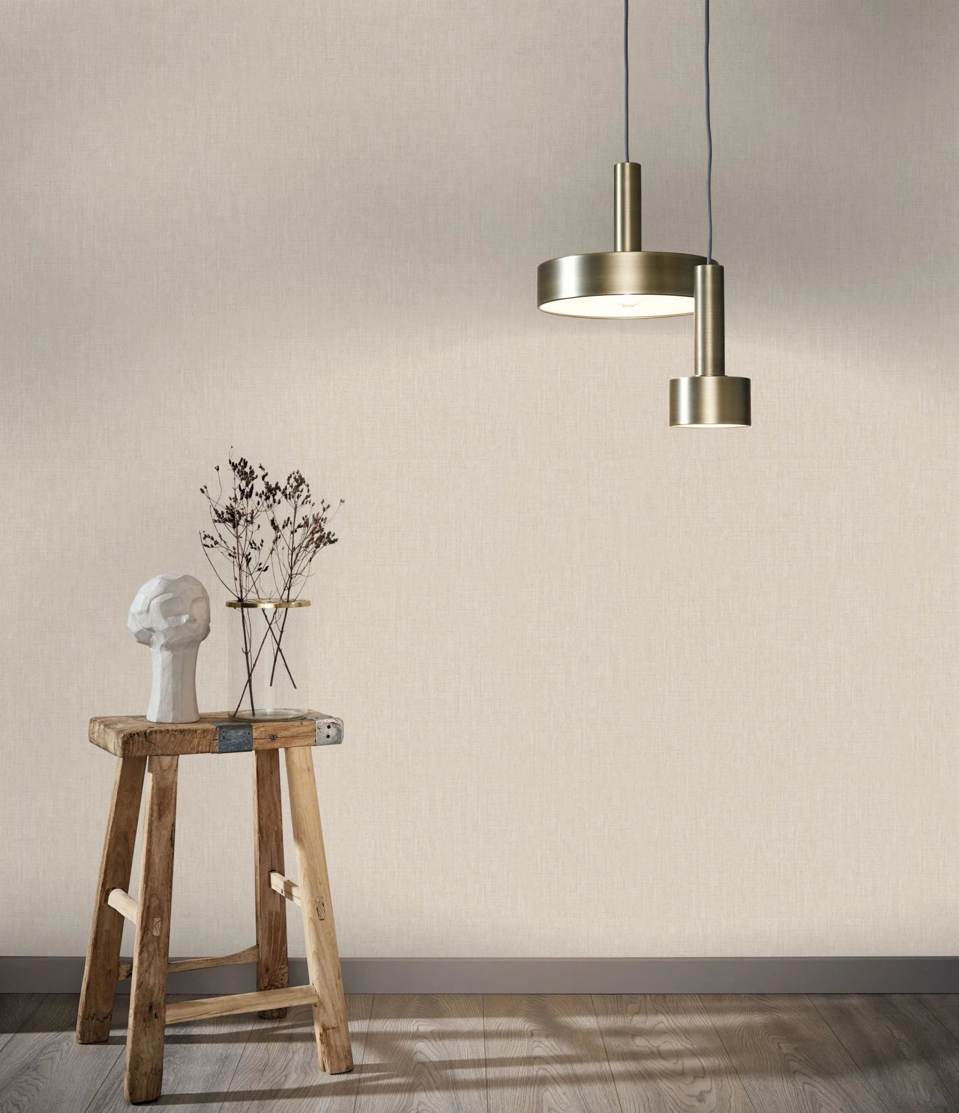 Marburg Botanica 33327 Light beige fabric-look wallpaper shown with two brass pendant lights and a rustic wooden stool holding an abstract white sculpture and a minimal vase with dried stems; wood floor and baseboard visible.