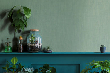 Marburg Botanica 33331 Green textured wallpaper behind a teal shelf displaying a glass terrarium with a cork lid, a plant mister, a glass bottle with a plant cutting, an air plant, and a small potted cactus with surrounding foliage.