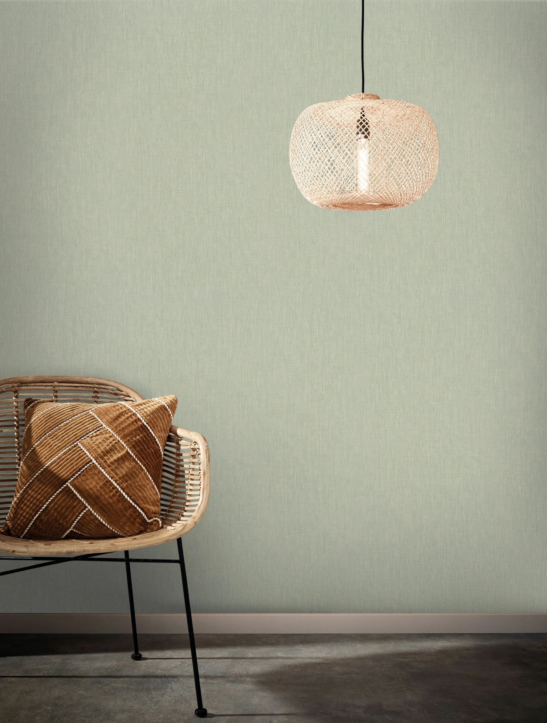 Marburg Botanica 33331 Soft green linen-texture wallpaper shown as a feature wall, styled with a rattan chair holding a brown cushion and a woven pendant light above.