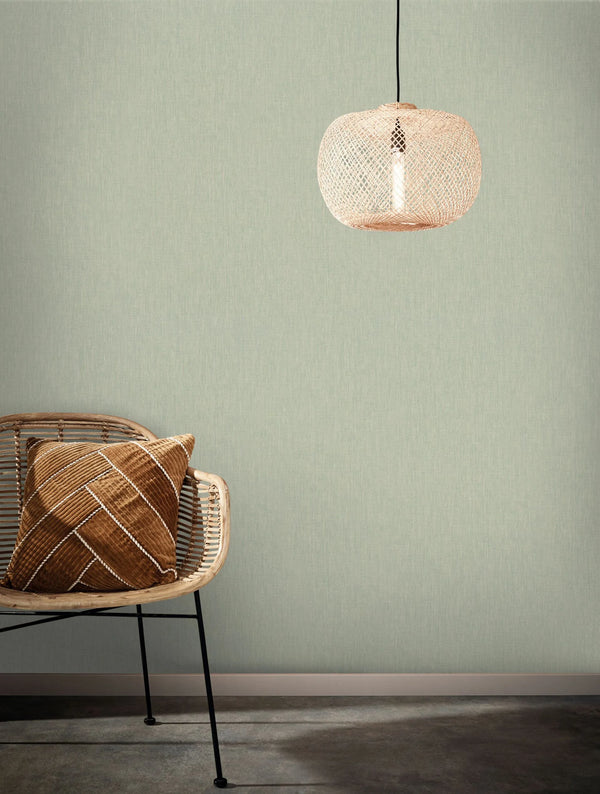 Marburg Botanica 33331 Soft green linen-texture wallpaper shown as a feature wall, styled with a rattan chair holding a brown cushion and a woven pendant light above.