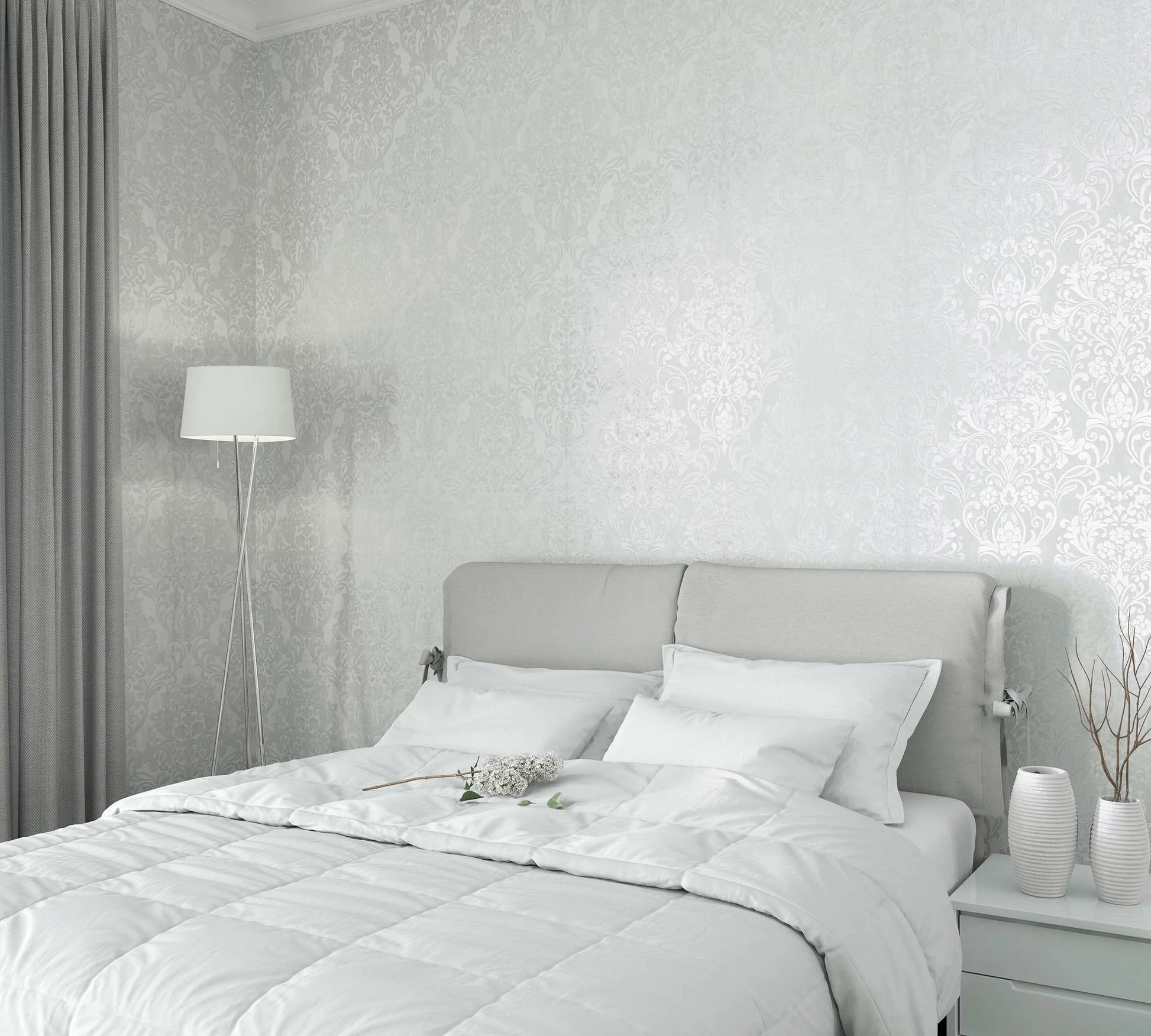Marburg City Glam 32601 Bedroom scene featuring light gray damask wallpaper with a soft sheen behind a light upholstered headboard, white bedding, a tripod floor lamp, gray curtains, and a nightstand with two ribbed vases and twig stems.