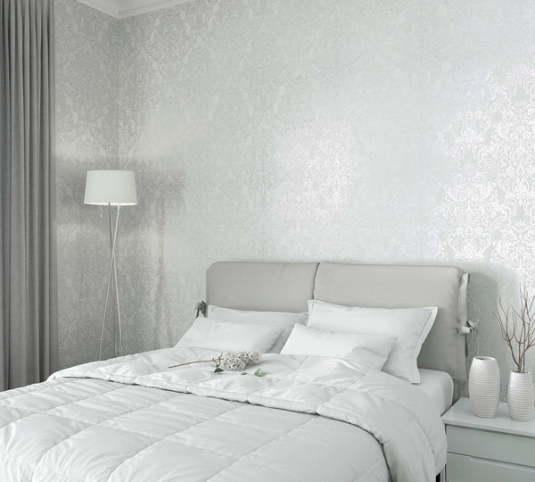 Marburg City Glam 32601 Bedroom scene featuring light gray damask wallpaper with a soft sheen behind a light upholstered headboard, white bedding, a tripod floor lamp, gray curtains, and a nightstand with two ribbed vases and twig stems.